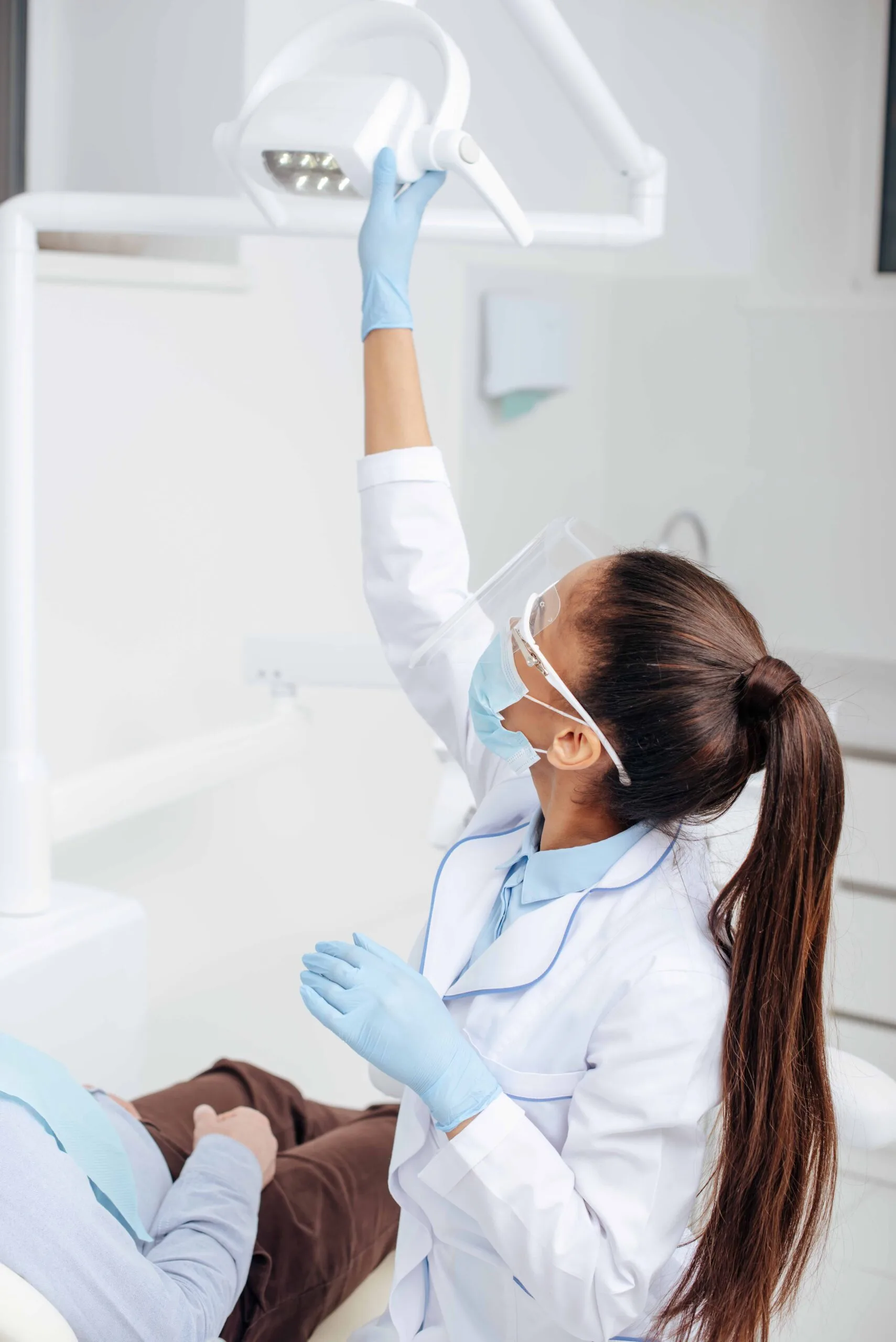 A dental professional adjusts an overhead light while wearing protective gear, including a mask, gloves, and eyewear. A patient is seated in the dental chair.