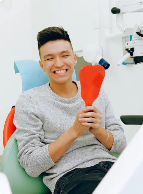Dr.-Langton-Dr-1 A smiling man in a dental chair holding a red hand mirror, wearing a gray sweater and black pants, in a bright dental office.