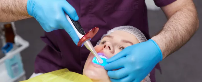 A dentist wearing blue gloves uses a curing light to harden dental sealant in a patient's mouth while they lie back in a dental chair, covered with a yellow protective sheet.