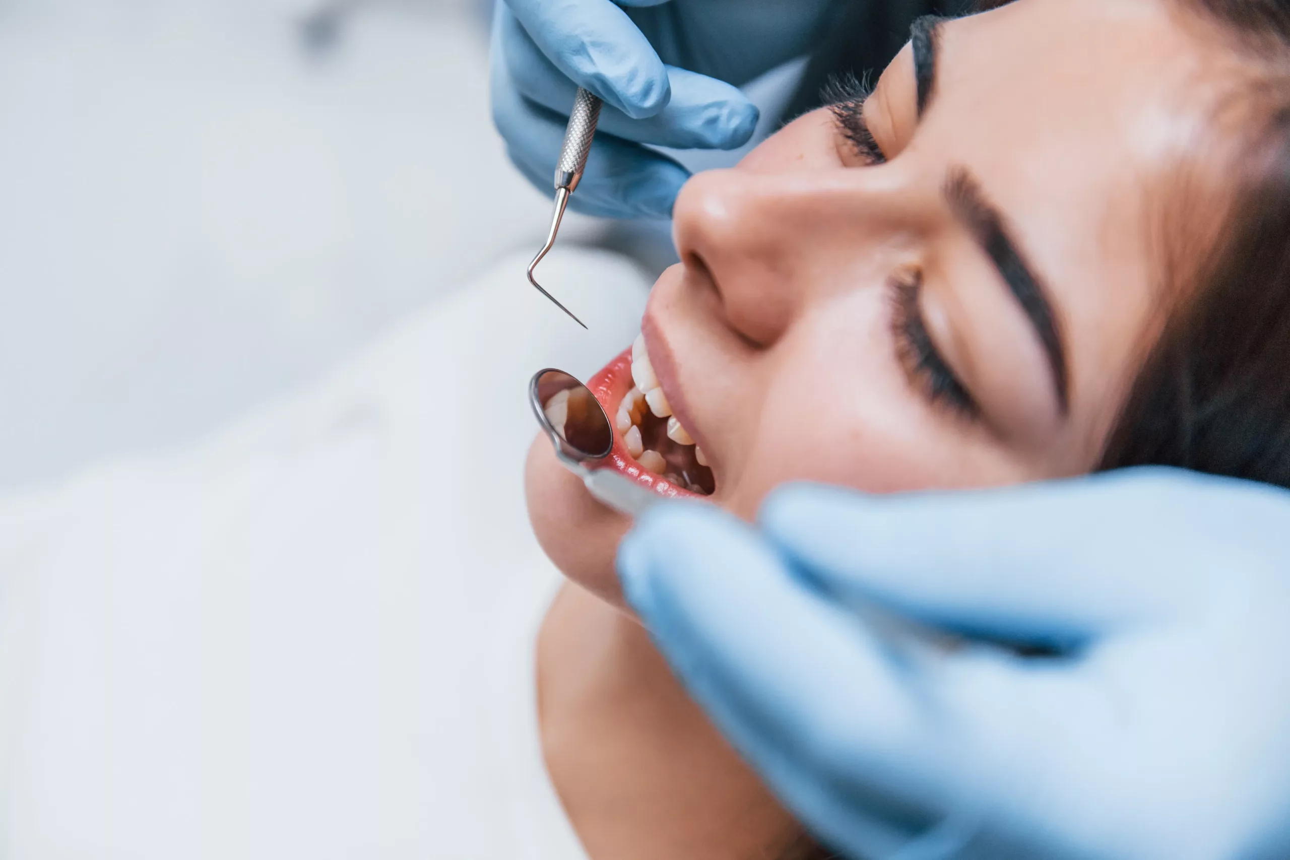 A dentist examines a patient's teeth using a dental mirror and probe, wearing blue gloves.
