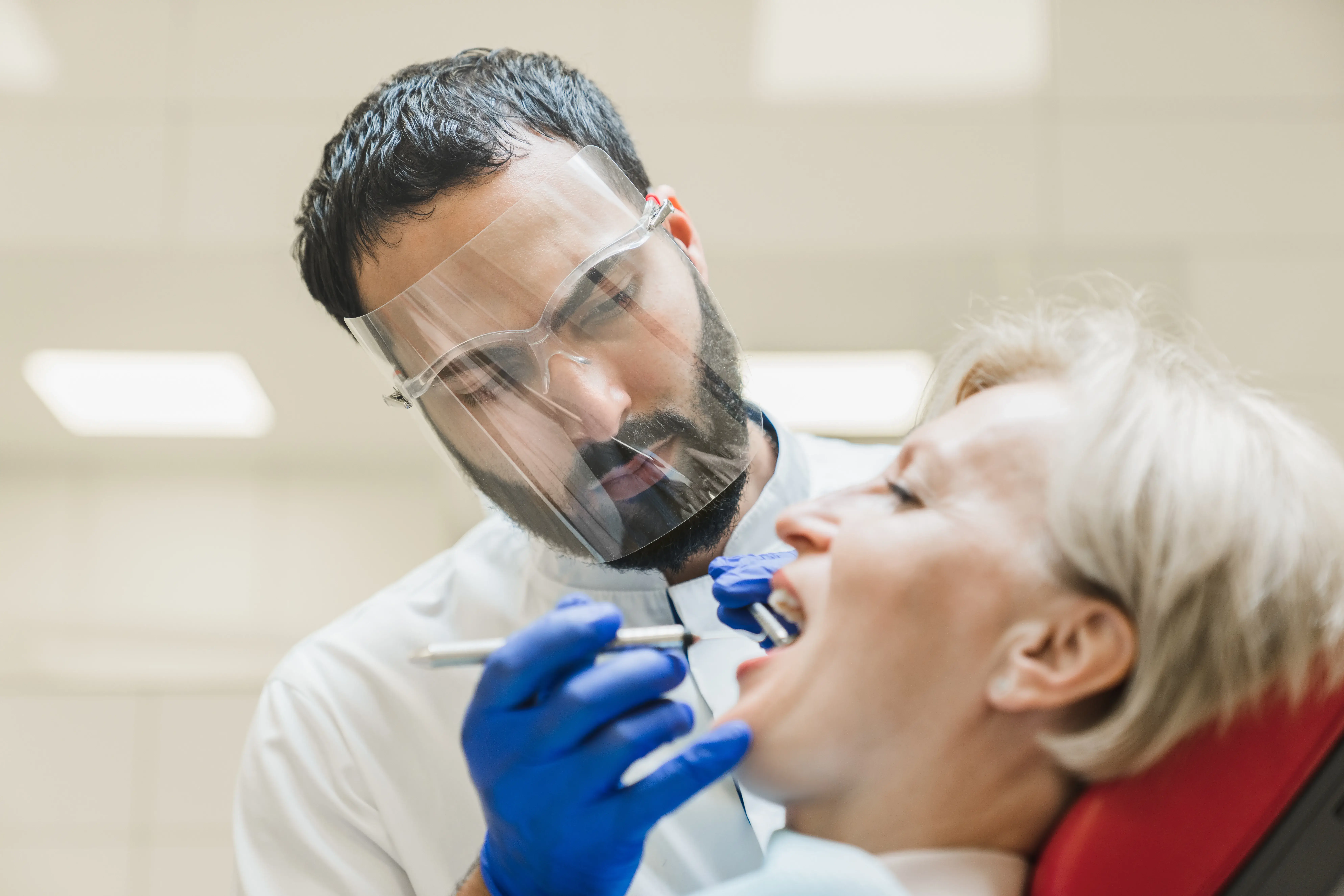 A dentist wearing protective gear examines a patient's teeth using dental tools.