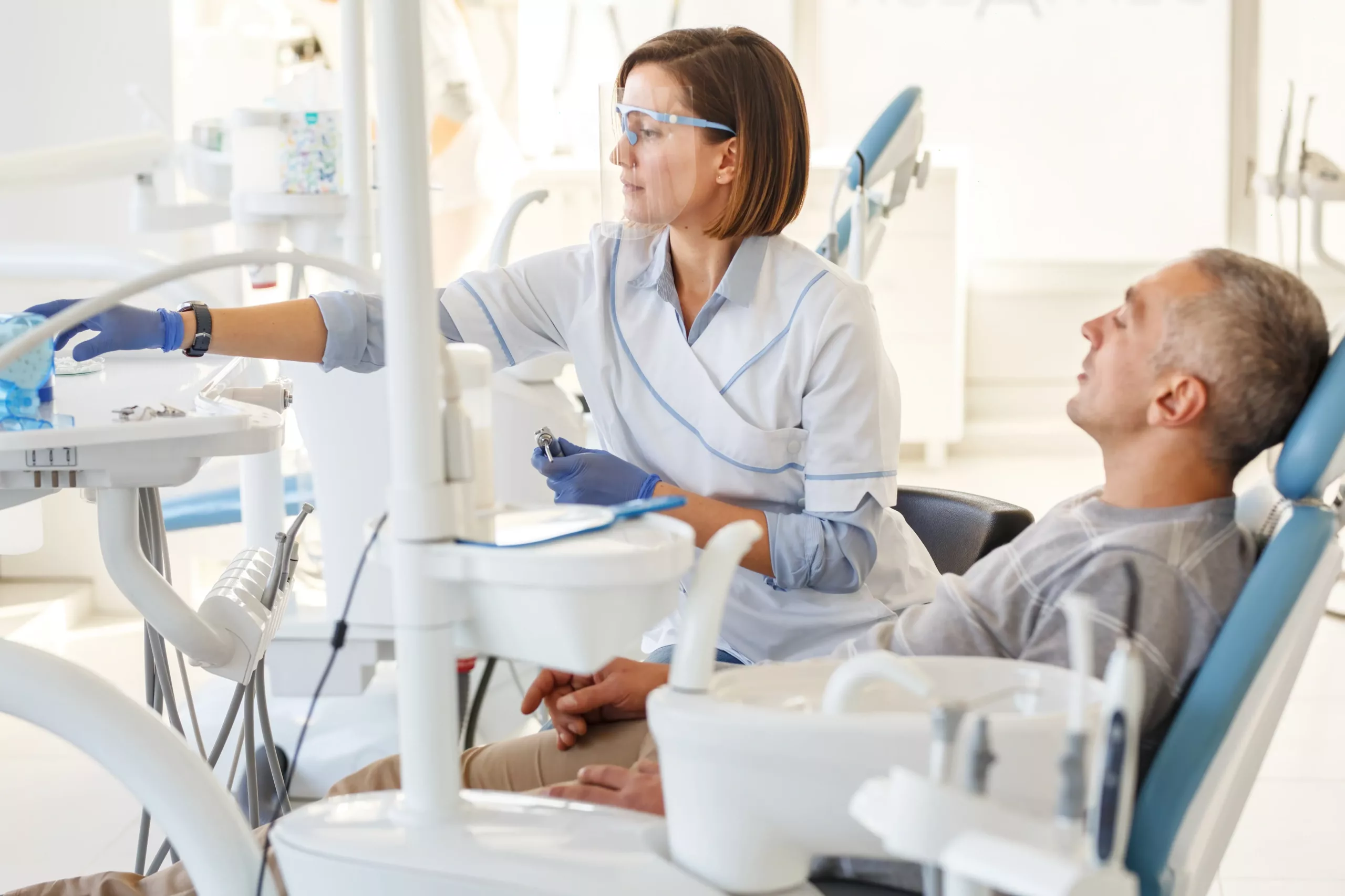 A dentist in protective eyewear and gloves prepares dental tools while a male patient sits in the chair in a dental office.