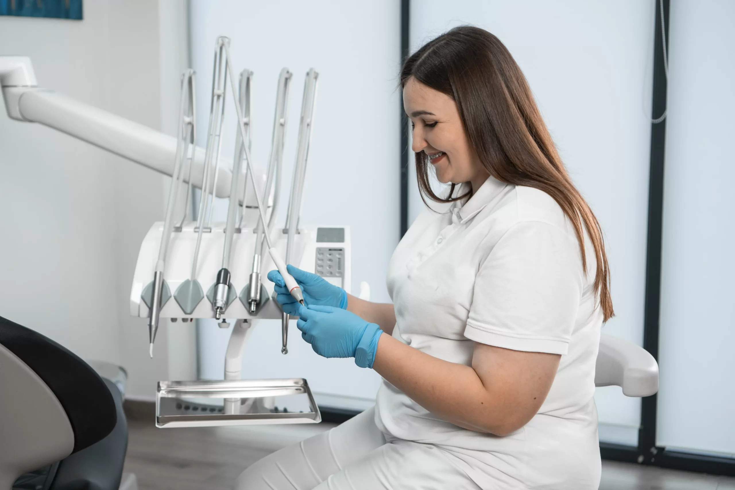A dental professional wearing gloves holds a tool while seated next to dental equipment, smiling in a modern office.