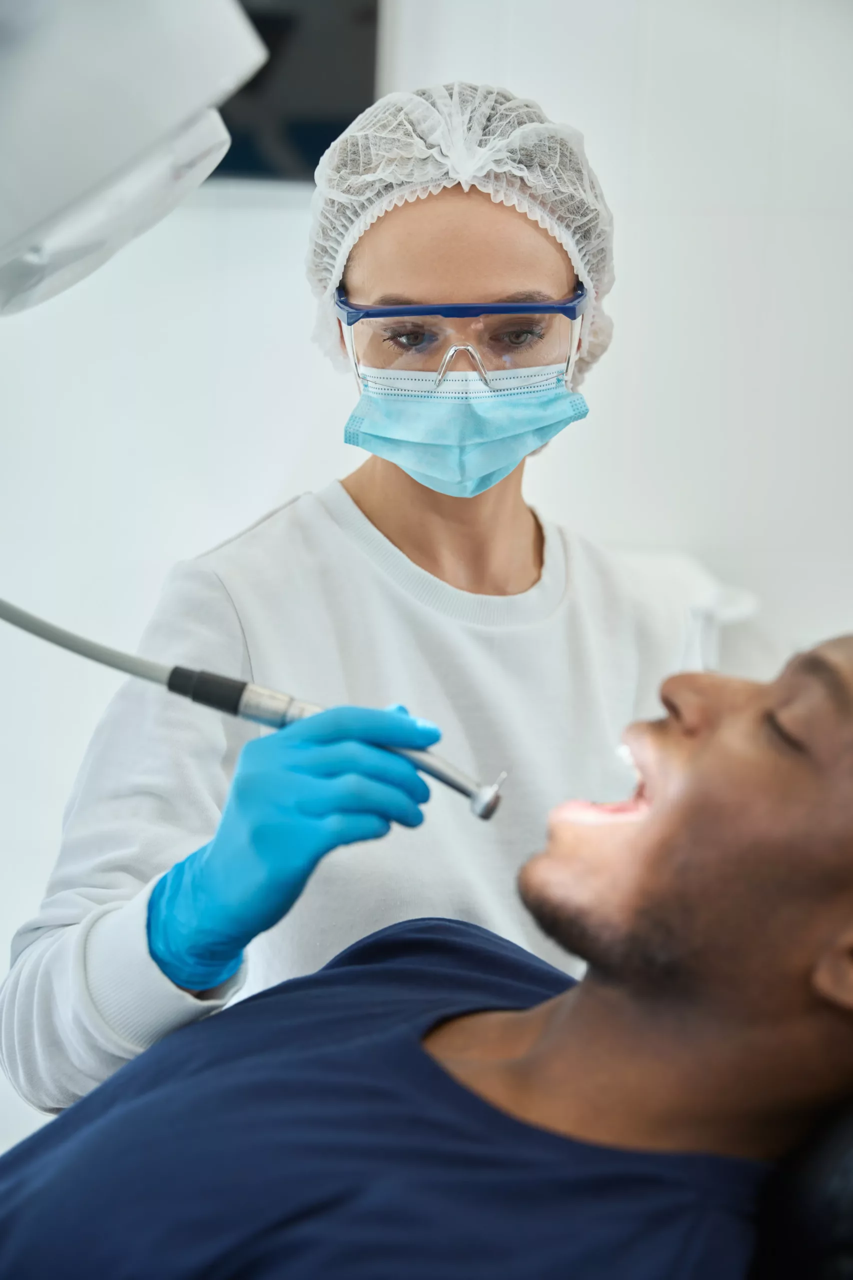 Dentist wearing protective gear examines a patient using dental tools. The patient is reclining with their mouth open.