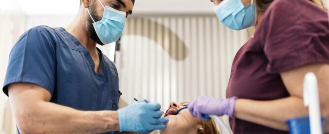 Two dental professionals wearing masks and scrubs perform a procedure on a patient lying in a dental chair.