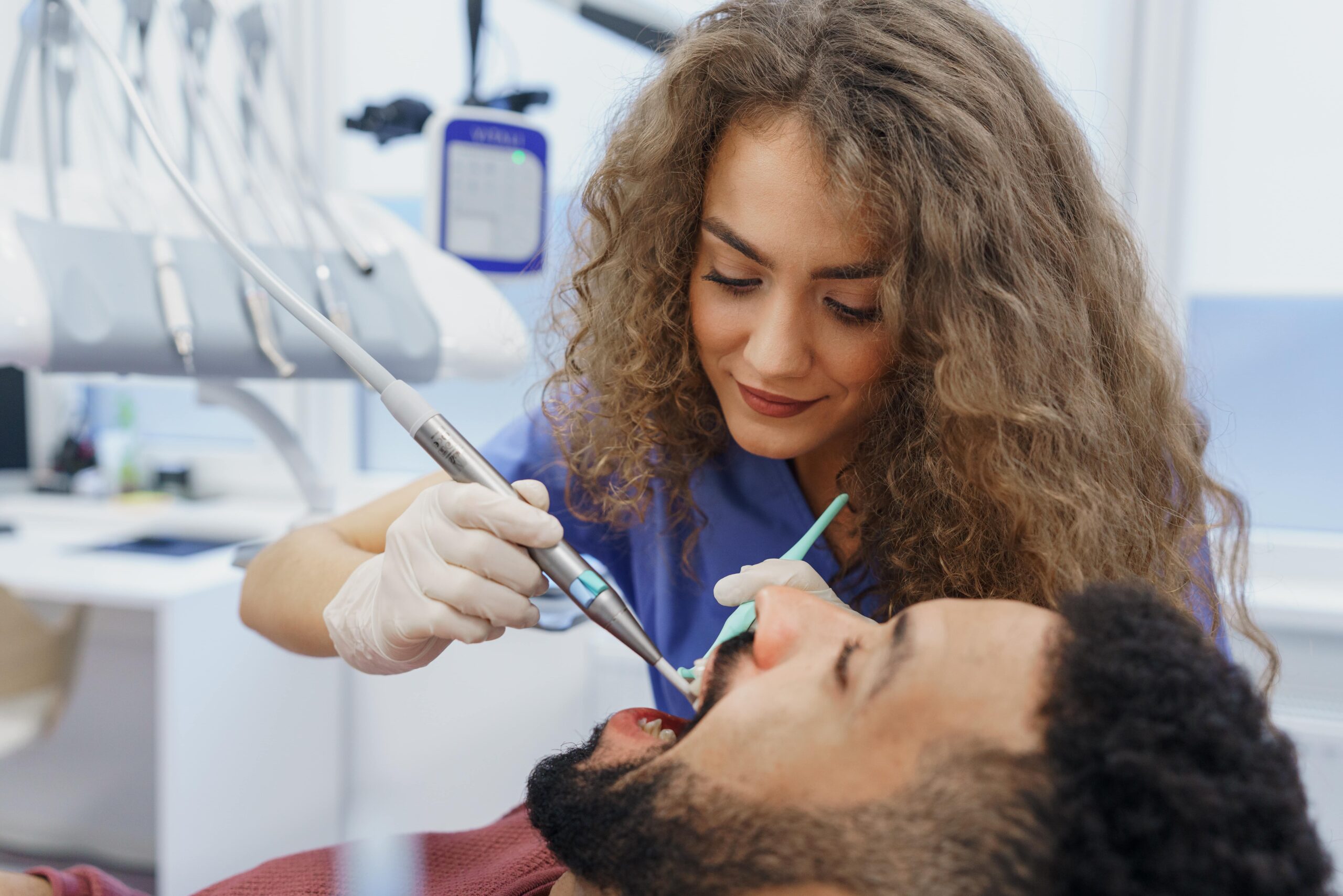 Dentist in blue scrubs using a dental drill and suction tool on a patient reclining in a dental chair, in a clinic setting.