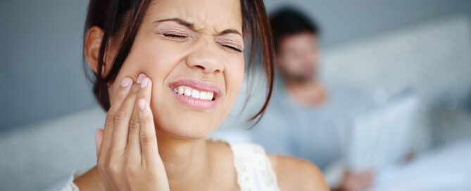 A woman sits on a bed, clutching her jaw with a pained expression, from teeth grinding a form of bruxism